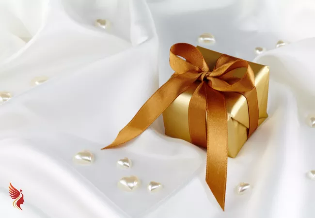 Geschenk in goldenem Papier mit goldroter Schleife auf einer weißen Decke trapiert mit perlmutt-farbenen Herzchen